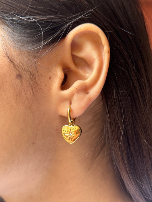 Gold heart-shaped earring worn on an ear with hair partially covering the frame.